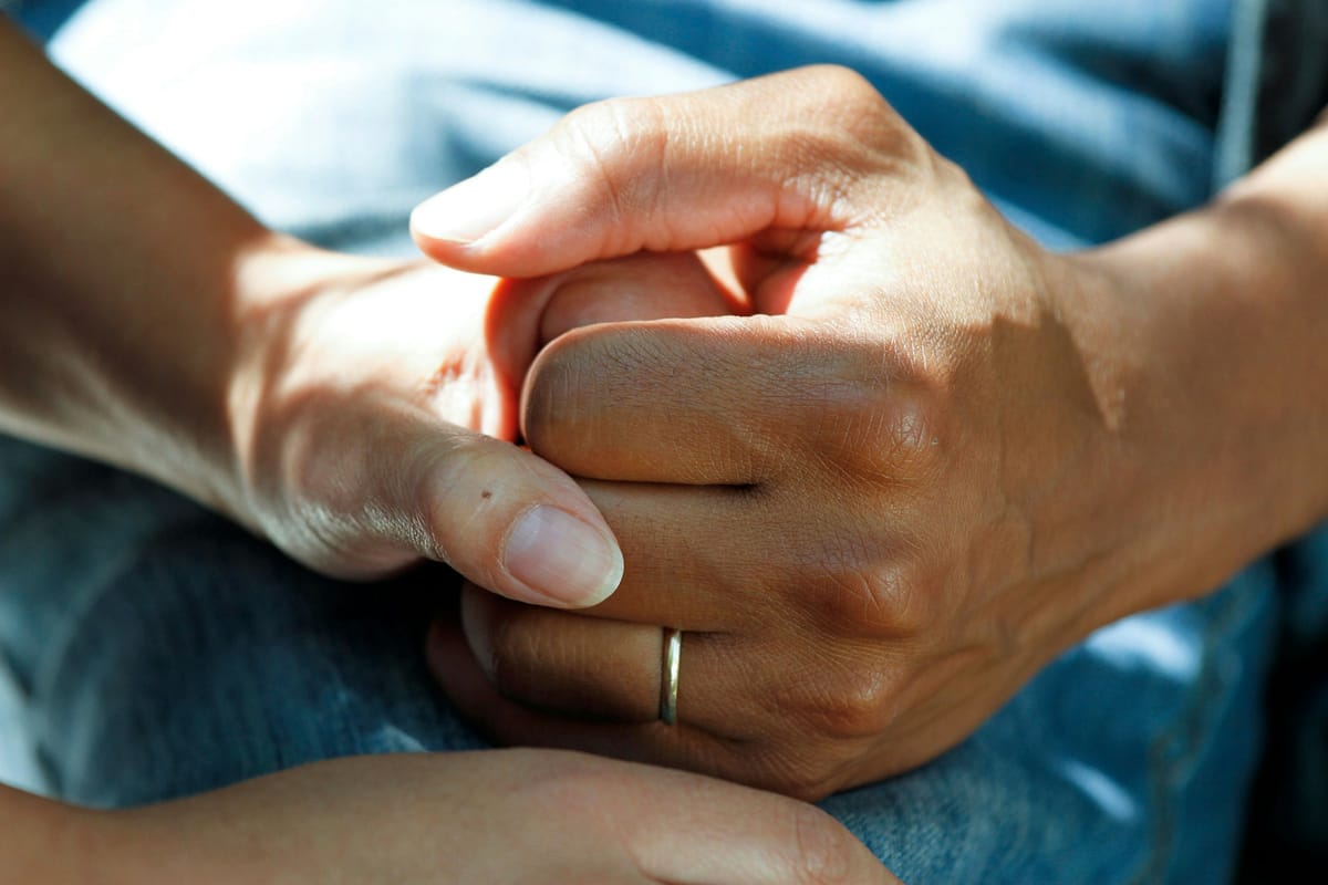 A close view of two people's hands clasped together over a lap in soft daylight, one hand resting over the other in a gesture of reassurance. No faces are in the frame.
