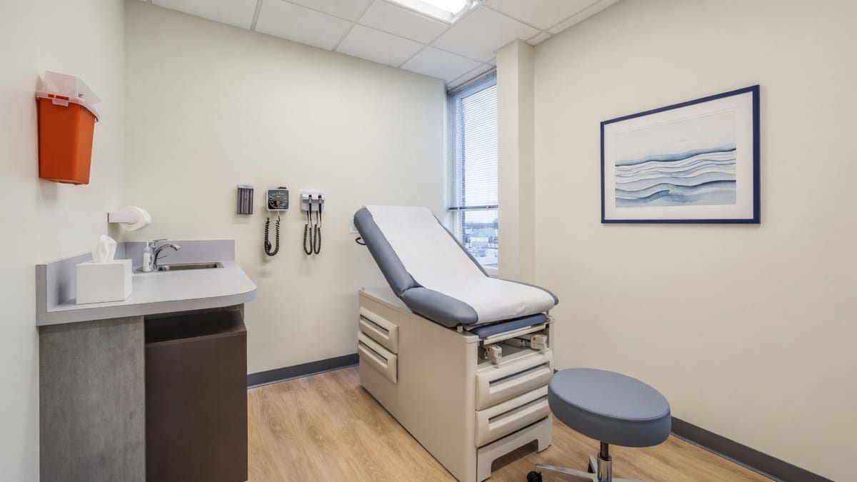 A small cream-walled exam room with pale oak flooring, a periwinkle-grey exam table with white paper, a round stool, a sink, wall-mounted diagnostic tools, a window with blinds, and a framed watercolor of blue water ripples.