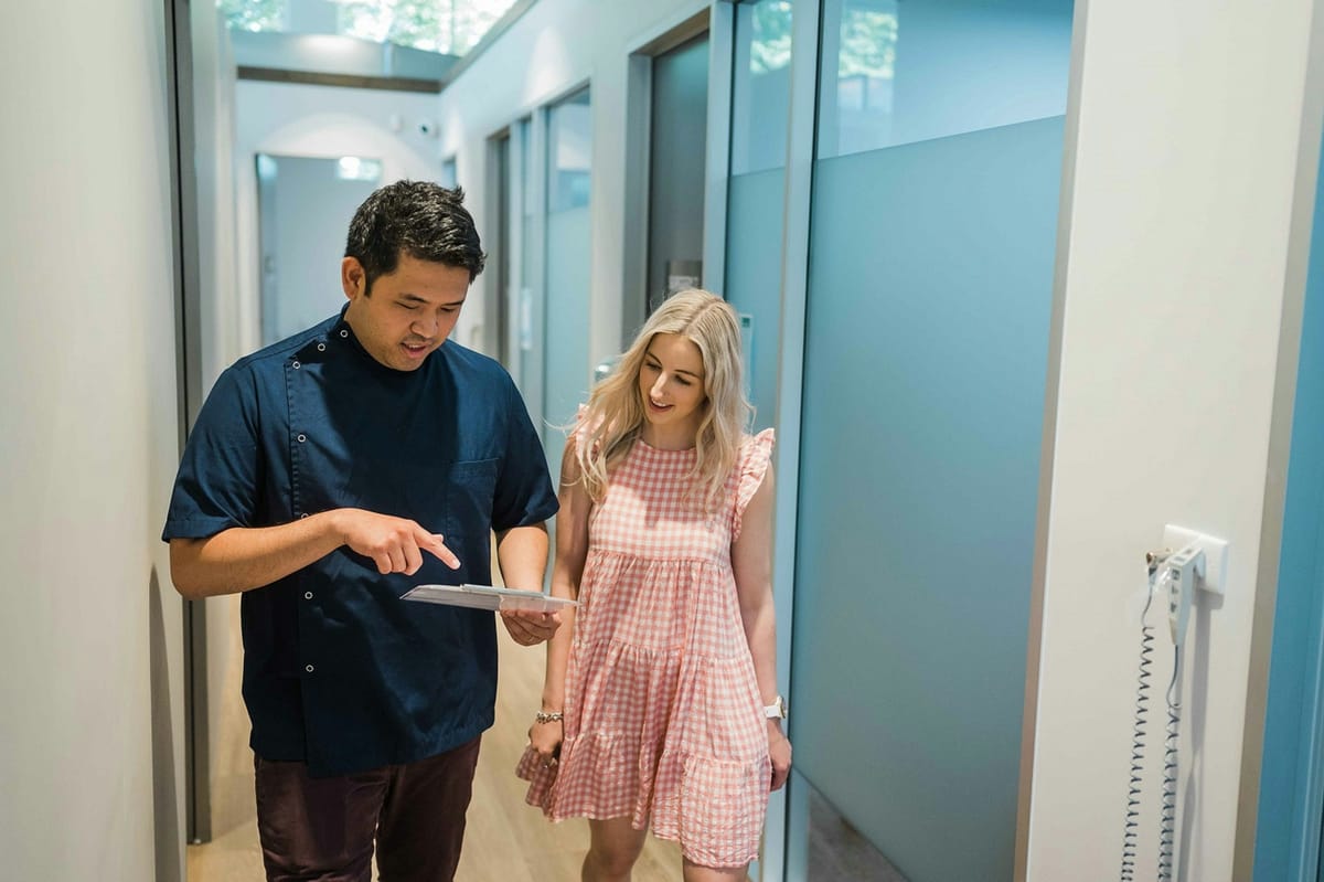 A clinician in a navy tunic walks a smiling patient in a pink gingham dress out along a bright, skylit clinic corridor, pointing at a small leaflet he is holding as they go.