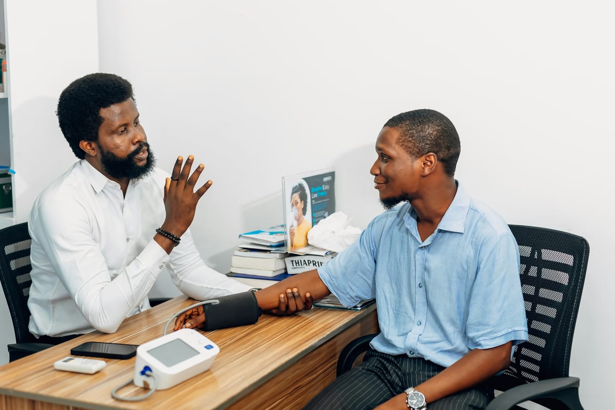 A man in a white shirt sits across a wooden desk from a man in a pale blue shirt, gesturing as he explains something; the seated man has a blood pressure cuff on his arm and the monitor rests on the desk between them.