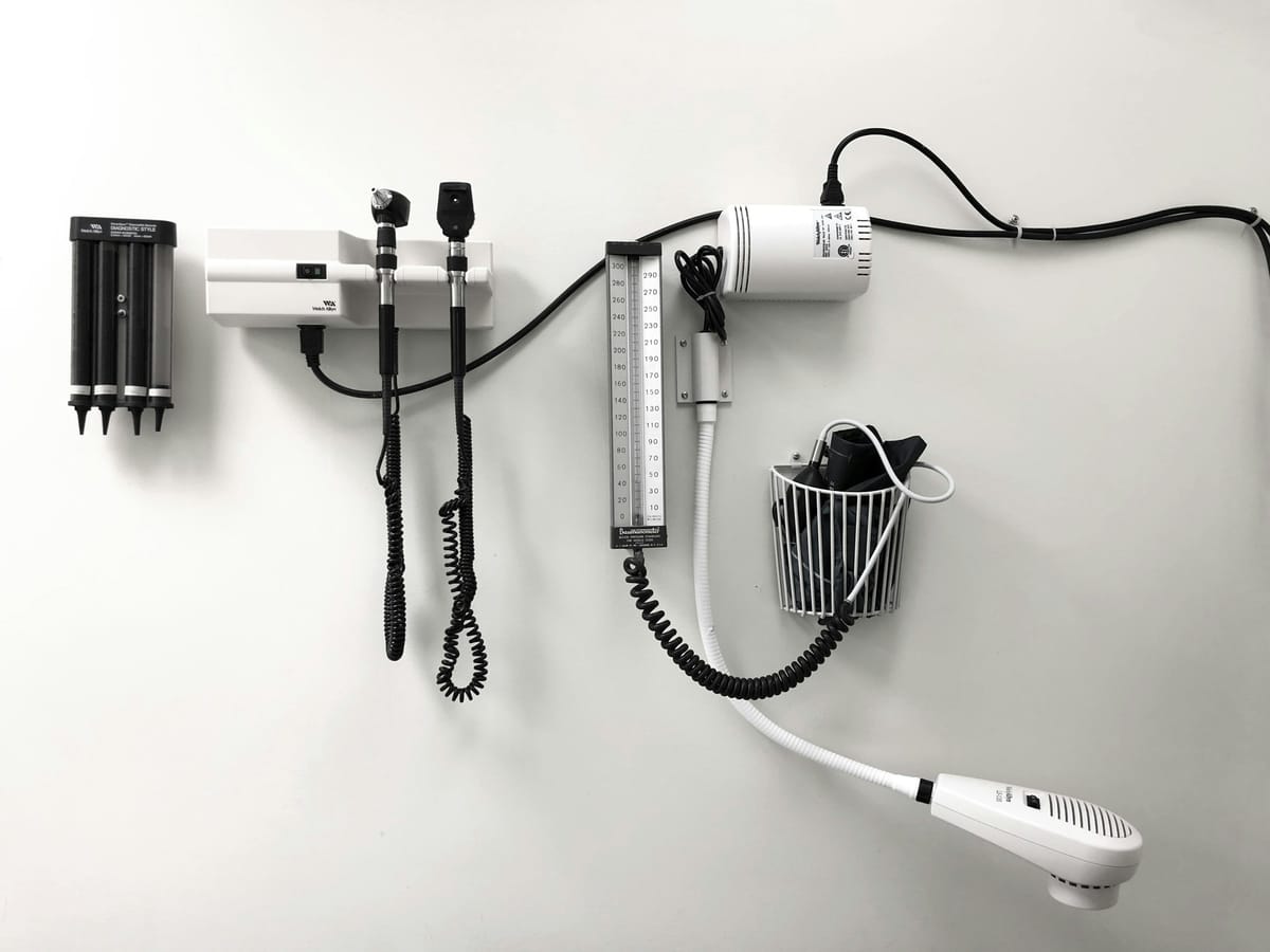 A white examination-room wall with wall-mounted diagnostic instruments: an otoscope and ophthalmoscope set with coiled black cords, a blood-pressure gauge, and a row of specula in a holder.