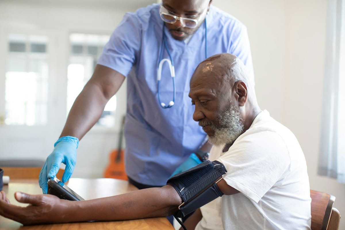 An older Black man in a white t-shirt sits at a wooden dining table with a blood-pressure cuff fastened around his upper arm, looking down at the monitor. A younger male clinician in pale blue scrubs, glasses and a stethoscope leans in beside him and steadies the monitor with a gloved hand. Behind them a bright window and a guitar are softly out of focus.
