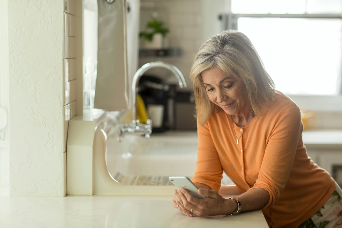 An older woman with shoulder-length blonde-grey hair, wearing an orange cardigan, leans on a white kitchen counter and smiles down at the smartphone held in both hands. Behind her a sink, tap and coffee machine sit softly out of focus in warm daylight from a window.